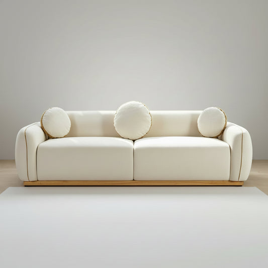 A cream-colored upholstered sofa with three round matching pillows, set in a bright modern living room with a marble coffee table, sheer curtains, and a large window overlooking green mountains.